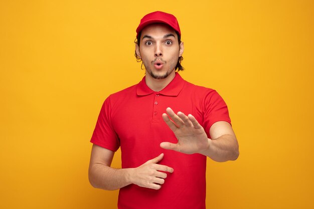 sorpreso giovane fattorino che indossa uniforme e cappuccio guardando la fotocamera che non mostra alcun gesto isolato su sfondo giallo