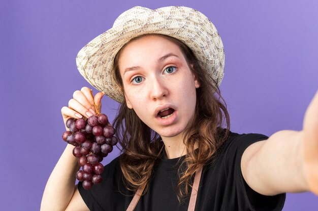 Sorpreso giovane giardiniere femminile che indossa cappello da giardinaggio tenendo l'uva con la fotocamera