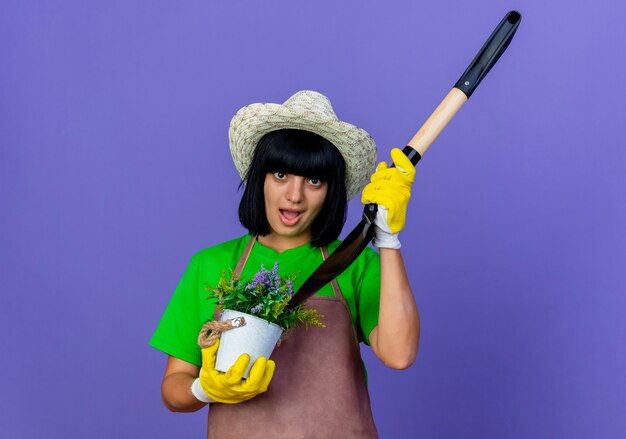 Sorpreso giovane giardiniere femminile in uniforme che indossa cappello e guanti da giardinaggio tiene la vanga