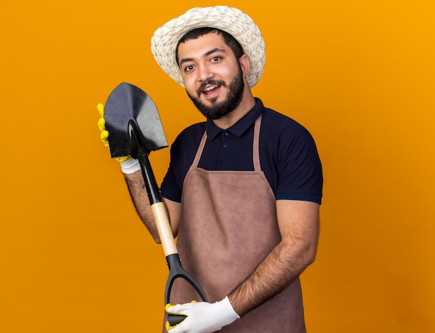 sorpreso giovane giardiniere maschio caucasico indossando guanti e cappello da giardinaggio in possesso di vanga isolato sulla parete arancione con spazio di copia