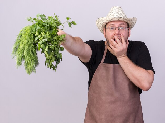 Sorpreso giovane giardiniere maschio che indossa cappello da giardinaggio tenendo fuori aneto con coriandolo alla telecamera isolata su muro bianco white