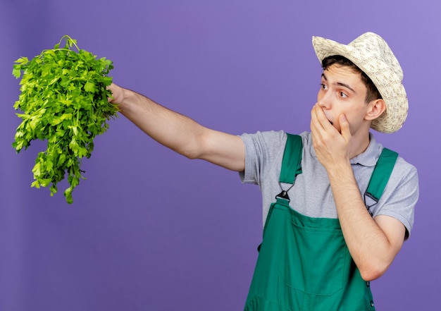 Sorpreso giovane giardiniere maschio che indossa il cappello da giardinaggio mette la mano sulla bocca tenendo il coriandolo