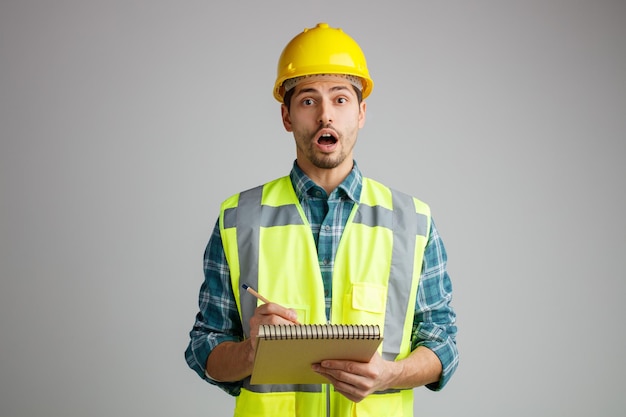 Sorpreso giovane ingegnere maschio che indossa casco di sicurezza e uniforme che tiene blocco note e matita guardando la fotocamera isolata su sfondo bianco