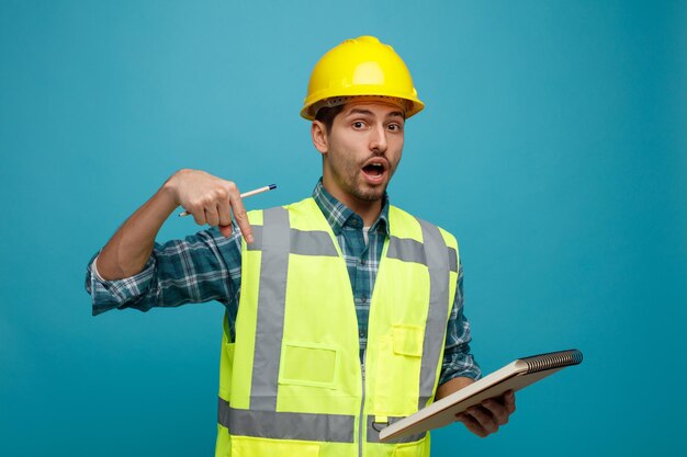 Sorpreso giovane ingegnere maschio che indossa casco di sicurezza e uniforme che tiene matita e blocco note guardando la fotocamera rivolta verso il basso isolata su sfondo blu