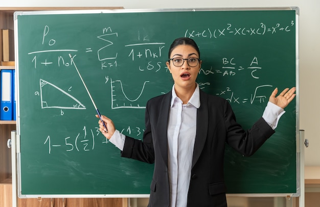Sorpreso giovane insegnante di sesso femminile con gli occhiali in piedi di fronte alla lavagna tenendo il puntatore stick allargando le mani in classe