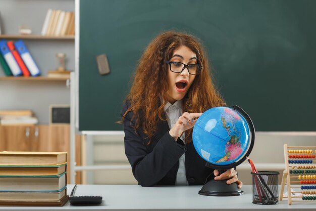 sorpreso giovane insegnante femminile che indossa occhiali in possesso e punta al globo seduto alla scrivania con strumenti scolastici in classe