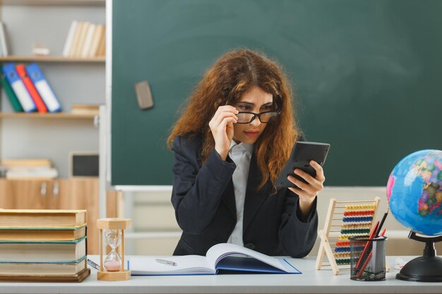 sorpreso giovane insegnante femminile che indossa occhiali tenendo e guardando la calcolatrice seduto alla scrivania con strumenti scolastici in classe