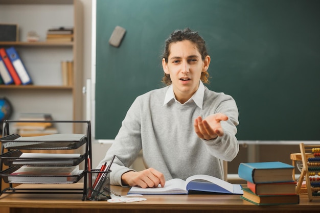 sorpreso giovane insegnante maschio che tiene la mano alla telecamera seduto alla scrivania con gli strumenti della scuola in classe