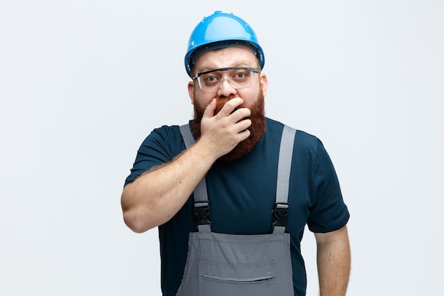 Sorpreso giovane lavoratore edile maschio che indossa l'uniforme del casco di sicurezza e occhiali di sicurezza tenendo la mano sulla bocca guardando la fotocamera isolata su sfondo bianco