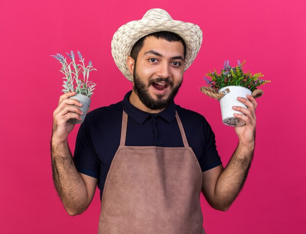 Sorpreso giovane maschio caucasico giardiniere che indossa cappello da giardinaggio tenendo vasi di fiori isolati sulla parete rosa con spazio di copia