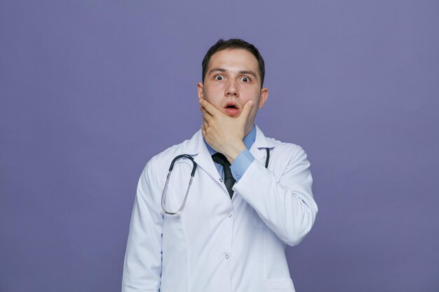 Sorpreso giovane medico maschio che indossa accappatoio medico e stetoscopio intorno al collo guardando la fotocamera tenendo la mano sul mento isolato su sfondo viola