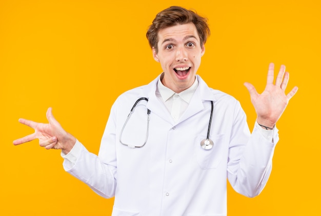 Sorpreso giovane medico maschio che indossa una tunica medica con uno stetoscopio che mostra numeri differenti isolati sulla parete arancione