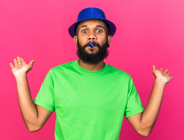 Sorpreso giovane ragazzo afro-americano che indossa il cappello da festa che soffia il fischio della festa allargando la mano isolata sul muro rosa