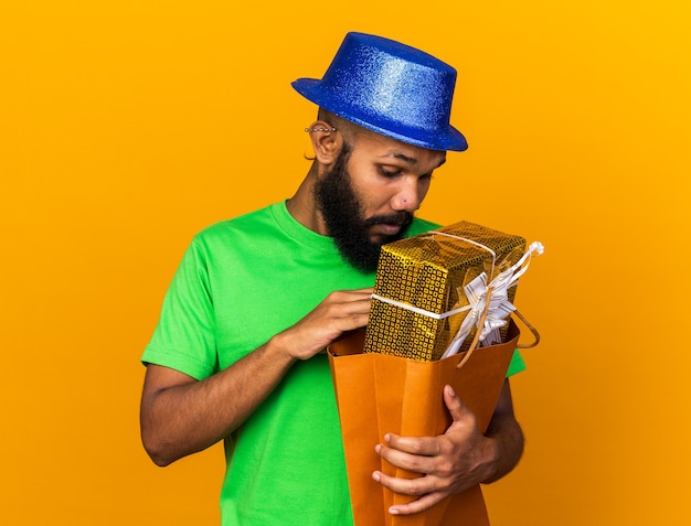 Sorpreso giovane ragazzo afroamericano che indossa un cappello da festa che tiene in mano e guarda nella borsa regalo