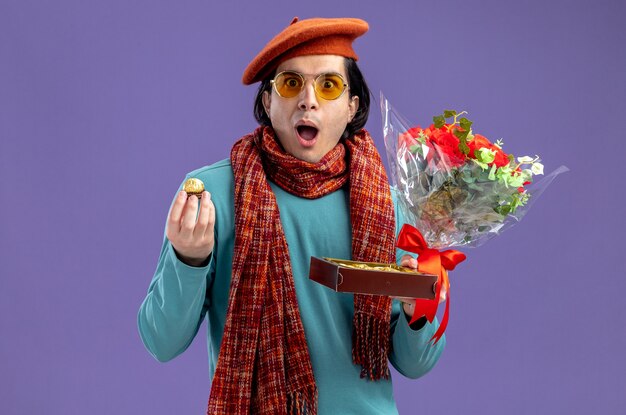 Sorpreso giovane ragazzo il giorno di San Valentino che indossa cappello con sciarpa e occhiali che tengono bouquet con scatola di caramelle isolato su sfondo blu