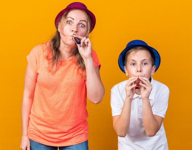 sorpreso giovane ragazzo slavo con cappello blu del partito e sua madre che indossa il cappello viola del partito che soffia fischietti del partito isolati sulla parete arancione con lo spazio della copia