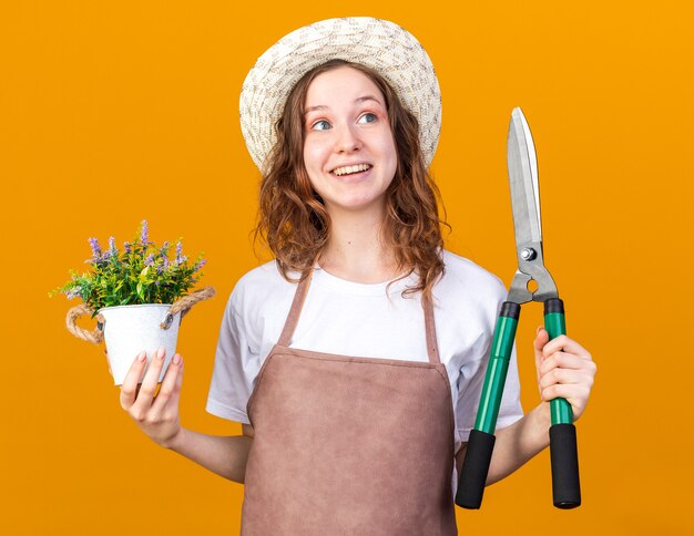 Sorpreso lato giovane giardiniere femminile che indossa cappello da giardinaggio tenendo il fiore in vaso di fiori con cesoie da potatura