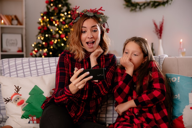 Sorpreso madre e figlia guardando il telefono seduto sul divano e godersi il periodo natalizio a casa