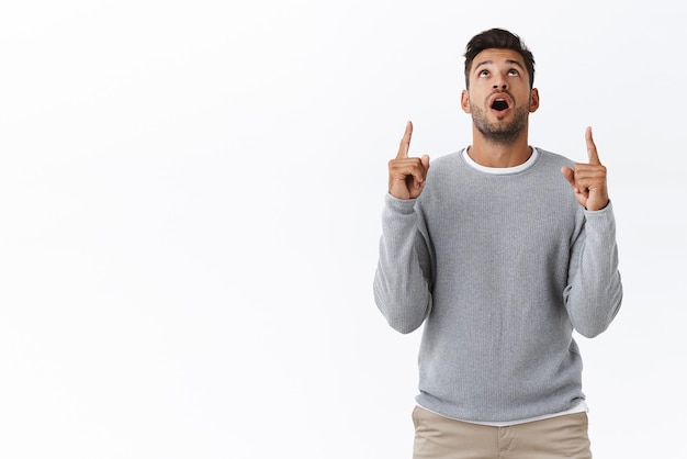 Sorpreso meravigliato bel ragazzo barbuto con taglio di capelli alla moda indossare casual maglione grigio goccia mascella impressionato alzare la testa e fissare verso l'alto dallo stupore rivolto verso l'alto con interesse e curiosità