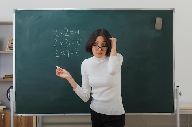 sorpreso mettendo la mano sulla testa giovane insegnante femminile con gli occhiali in piedi davanti alla lavagna e scrive in classe