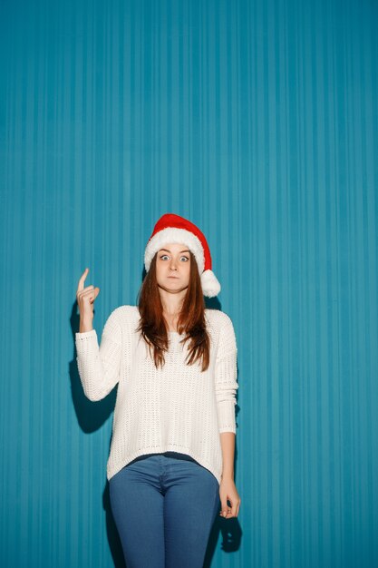sorpreso natale ragazza che indossa un cappello da Babbo Natale rivolto verso l'alto sullo sfondo blu studio
