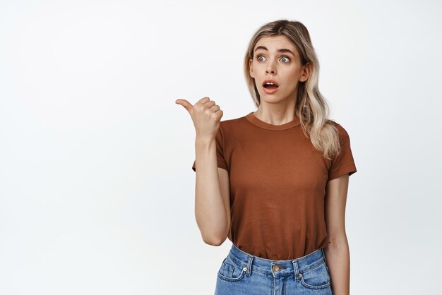 Sorpreso ragazza bionda sussulto impressionato dito puntato a sinistra e guardando la pubblicità stupito in piedi su sfondo bianco