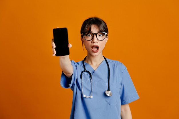 Sorpreso tenendo il telefono alla telecamera giovane dottoressa che indossa uno stetoscopio fith uniforme isolato su sfondo arancione