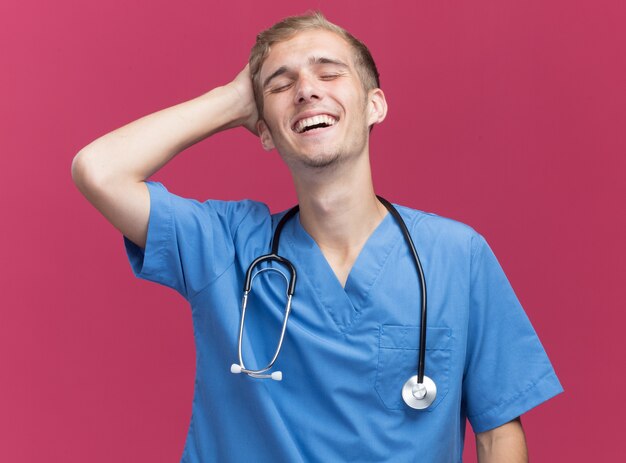 Sorridendo con gli occhi chiusi giovane medico maschio che indossa l'uniforme del medico con lo stetoscopio che mette la mano sulla testa isolata sulla parete rosa