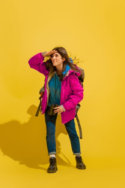 Sorridendo, sembra felice. Ritratto di una giovane ragazza turistica caucasica allegra con borsa e binocolo isolato su sfondo giallo studio. Prepararsi per il viaggio. Resort, emozioni umane, vacanze.