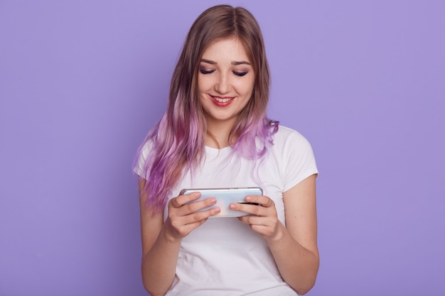 Sorridente abiti femminili maglietta bianca che tiene smart phone nelle mani, gioco di placcatura, guardando il display con un sorriso carino, utilizzando il telefono cellulare, in posa isolato sopra il muro viola.