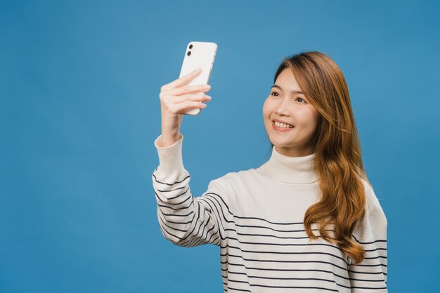 Sorridente adorabile femmina asiatica che fa foto selfie su smart phone con espressione positiva in abbigliamento casual e stand isolato su parete blu