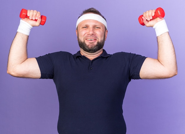 Sorridente adulto slavo sportivo uomo che indossa archetto e braccialetti che tengono i manubri isolati sulla parete viola con spazio di copia