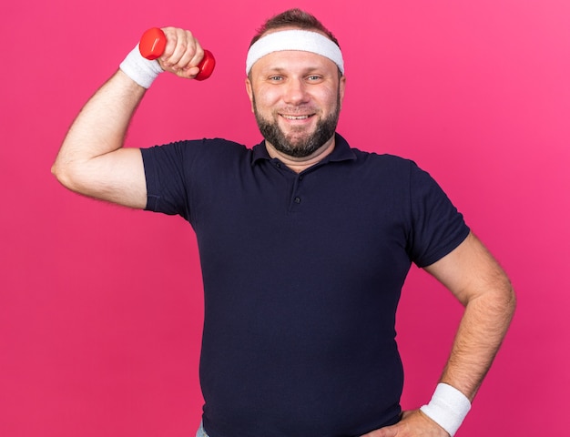 sorridente adulto slavo sportivo uomo che indossa la fascia e braccialetti tensing bicipite tenendo il manubrio isolato sul muro rosa con spazio di copia