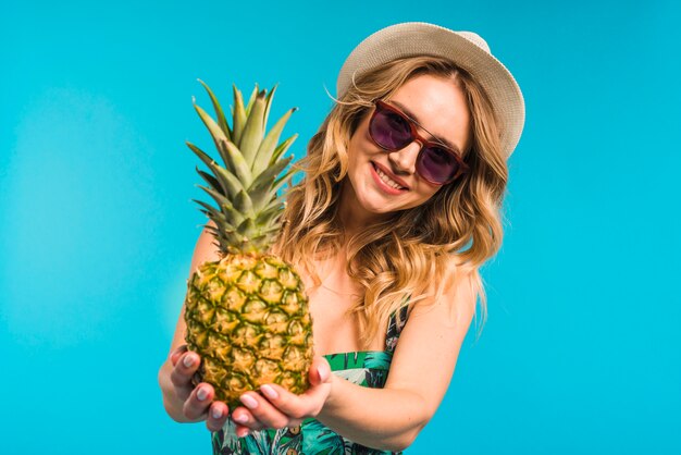 Sorridente attraente giovane donna in cappello e occhiali da sole in possesso di ananas fresco