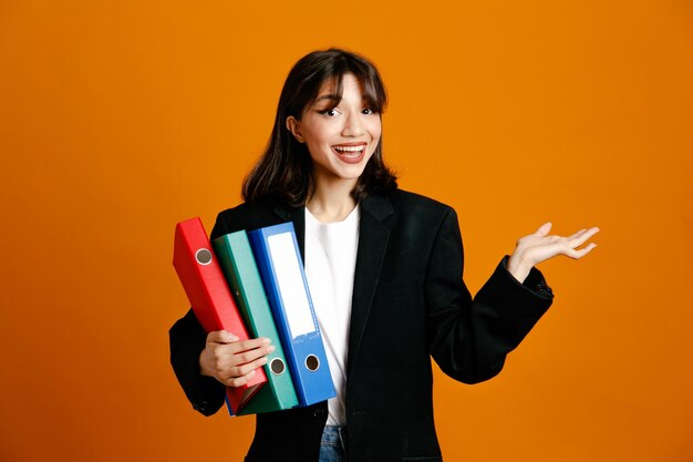 Sorridente azienda cartelle giovane bella femmina che indossa giacca nera isolata su sfondo arancione