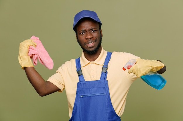 sorridente azienda detergente con straccio giovane maschio pulitore afroamericano in uniforme con guanti isolati su sfondo verde