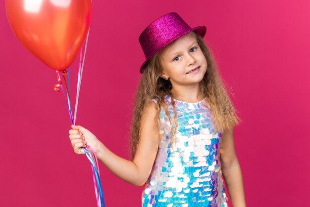 Sorridente bambina bionda con cappello da festa viola che tiene palloncini di elio e sembra isolata sulla parete rosa con spazio di copia copy