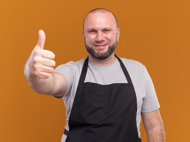 Sorridente barbiere maschio di mezza età in uniforme che mostra pollice in su isolato sulla parete arancione