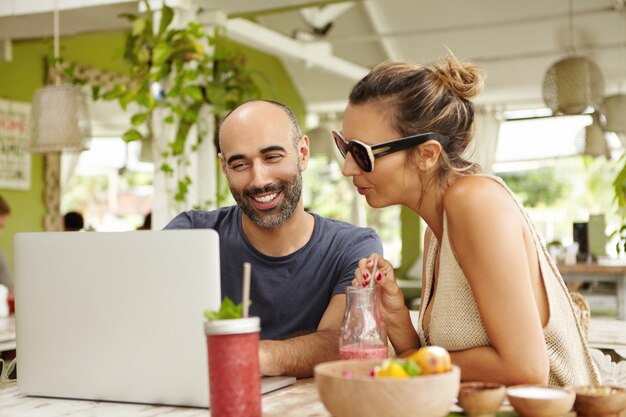 Sorridente barbuto uomo e donna in occhiali da sole seduti davanti al computer portatile aperto e discutendo di qualcosa, guardando lo schermo con interesse.