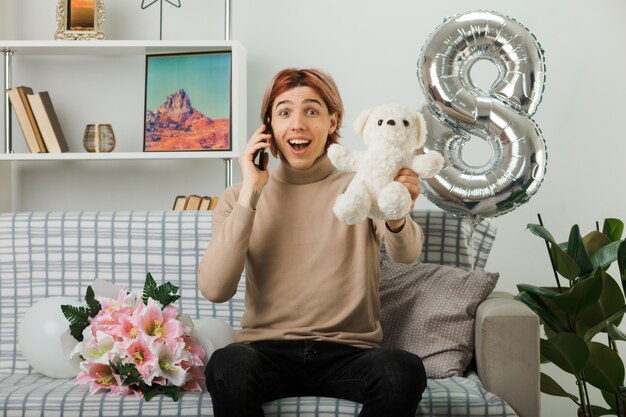 Sorridente bel ragazzo durante la giornata delle donne felici che porge orsacchiotto, parla al telefono seduto sul divano in soggiorno