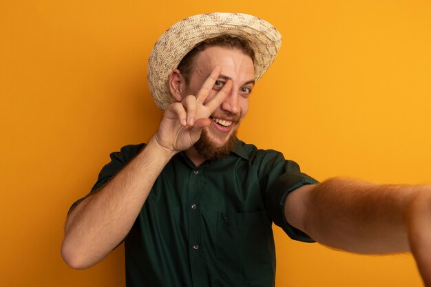 Sorridente bell'uomo biondo con cappello da spiaggia gesti il segno della mano di vittoria e finge di tenere la parte anteriore isolata sulla parete arancione