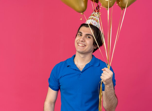Sorridente bell'uomo caucasico che indossa un berretto da compleanno tiene palloncini di elio