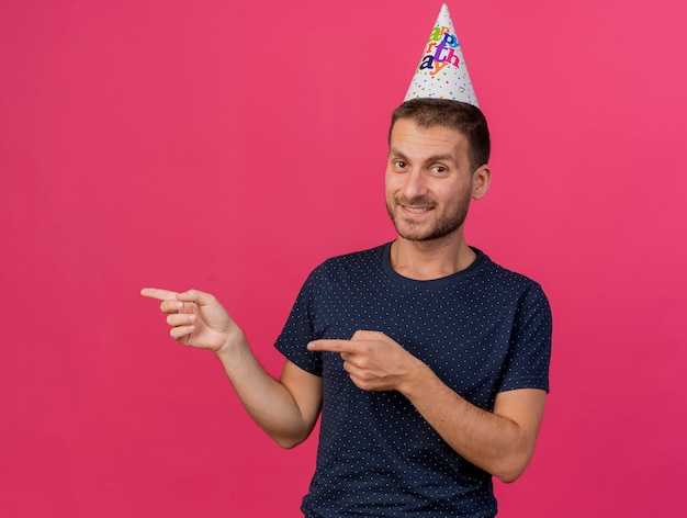 Sorridente bell'uomo caucasico indossando il cappello di compleanno che punta a lato con due mani isolate su sfondo rosa con spazio di copia