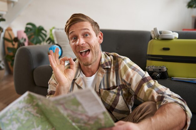 Sorridente bell'uomo turistico con una mappa stradale si siede sul pavimento vicino alla valigia mostra il segno ok ok