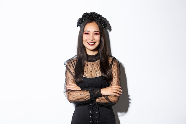 Sorridente bella donna asiatica che celebra halloween, indossa una corona nera e vestito, trucco gotico, guardando felice a porte chiuse. Femmina che celebra evento autunnale, dolcetto o scherzetto in costume.