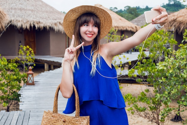 Sorridente bella donna che fa autoritratto dal telefono cellulare durante la sua vacanza tropicale in Thailandia. Vestiti blu luminosi estivi ^ cappello e borsa alla moda di paglia. Labbra rosse. Buonumore.