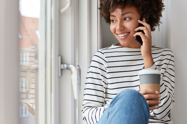 Sorridente bella donna dalla pelle scura parla sul dispositivo smart phone, detiene il caffè da asporto