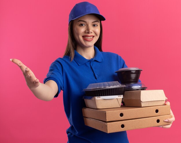 Sorridente bella donna di consegna in punti uniformi con la mano e tiene il pacchetto di cibo e contenitori su scatole per pizza isolato sulla parete rosa con spazio di copia