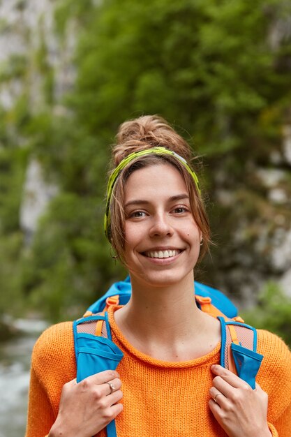 Sorridente bella donna viaggiatore in maglione arancione, porta zaino, indossa la fascia