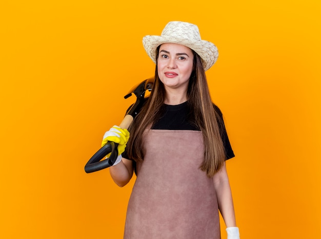 Sorridente bella ragazza giardiniere che indossa uniforme e cappello da giardinaggio con guanti mettendo vanga sulla spalla isolato su sfondo arancione